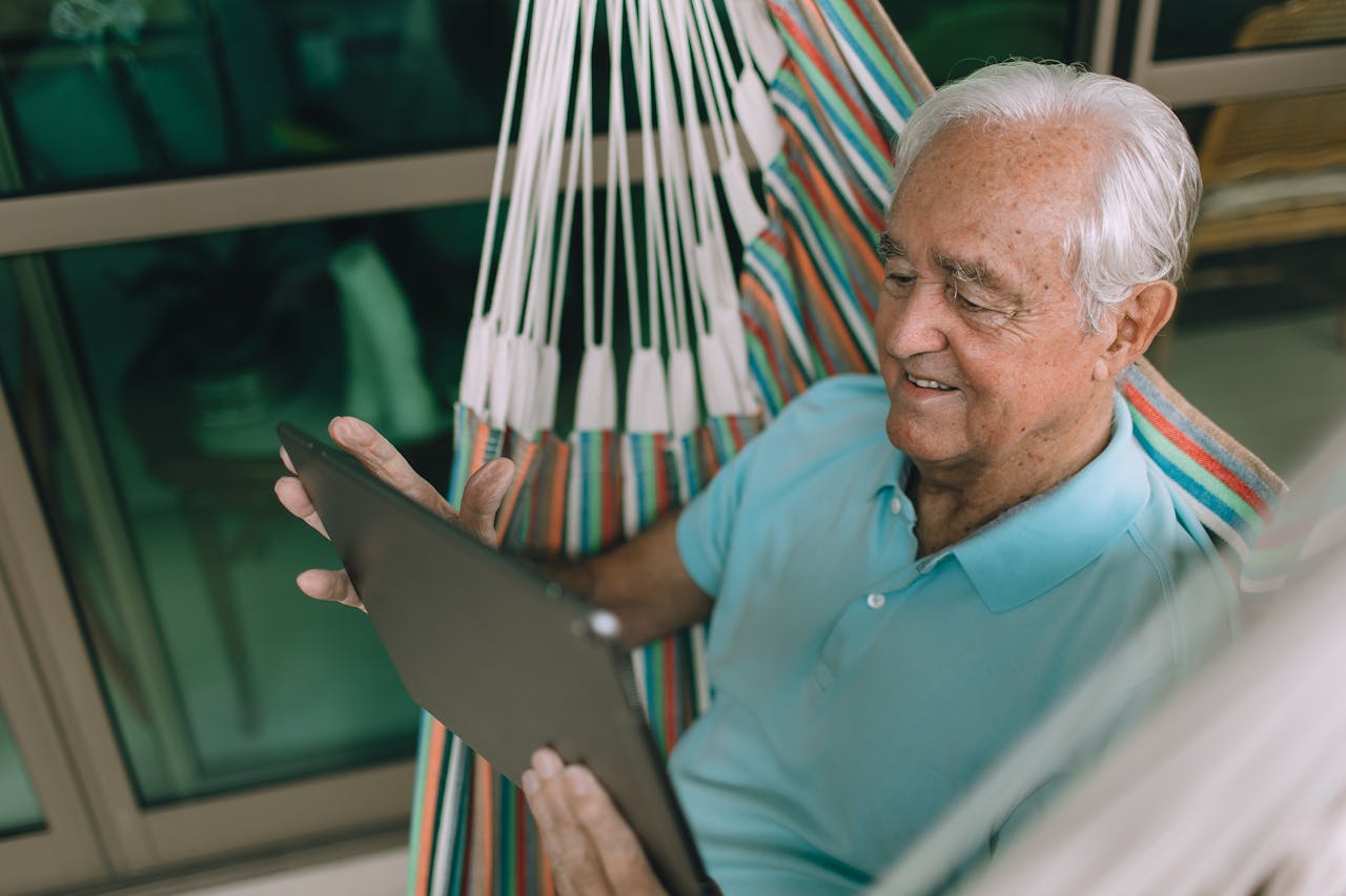 Senior man relaxing in a hammock indoors while using a tablet, enjoying leisure time.